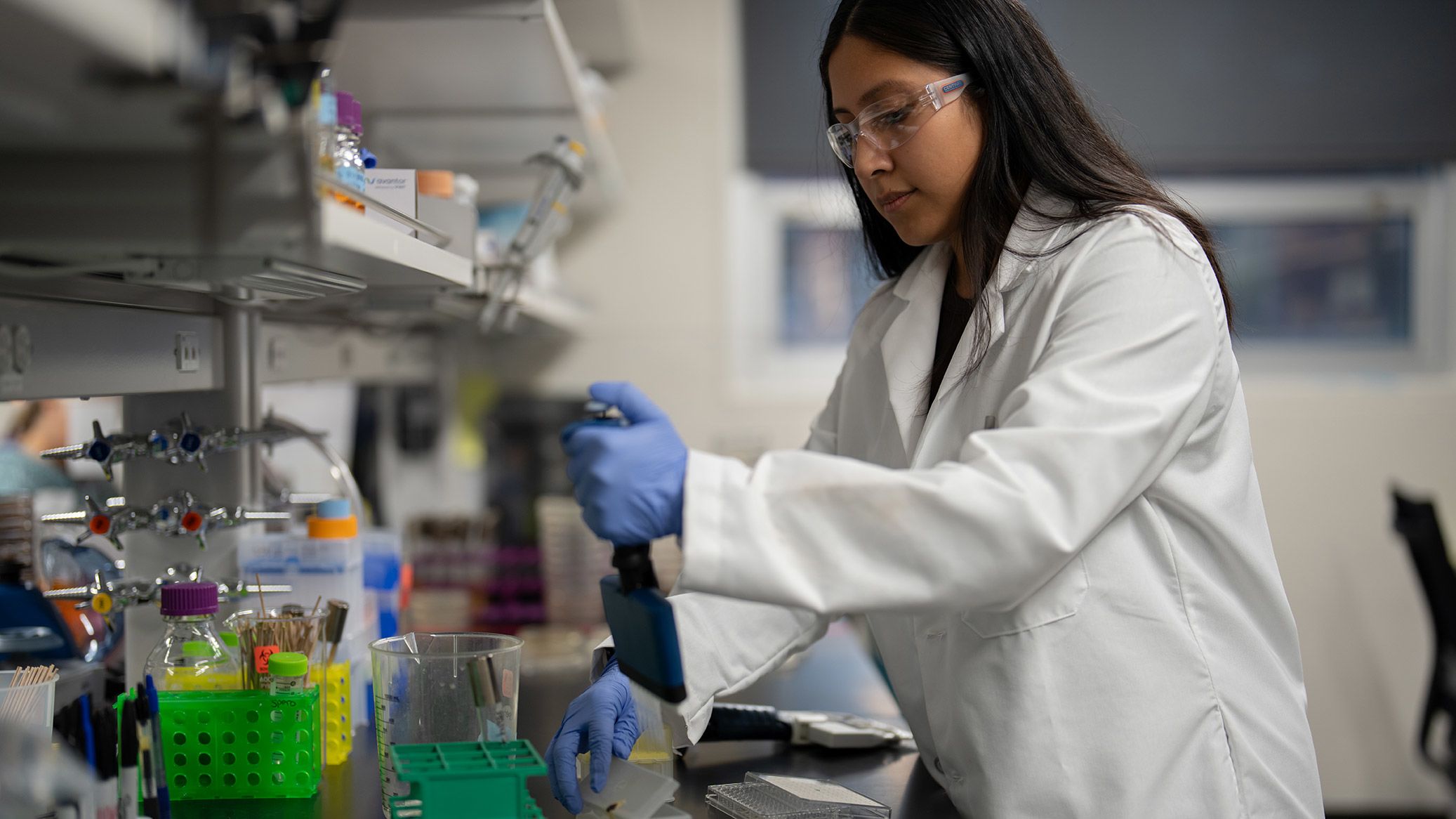 Celine Lopez Padilla wears a lab coat, safety glasses, and gloves and uses a pipette at a lab workbench.