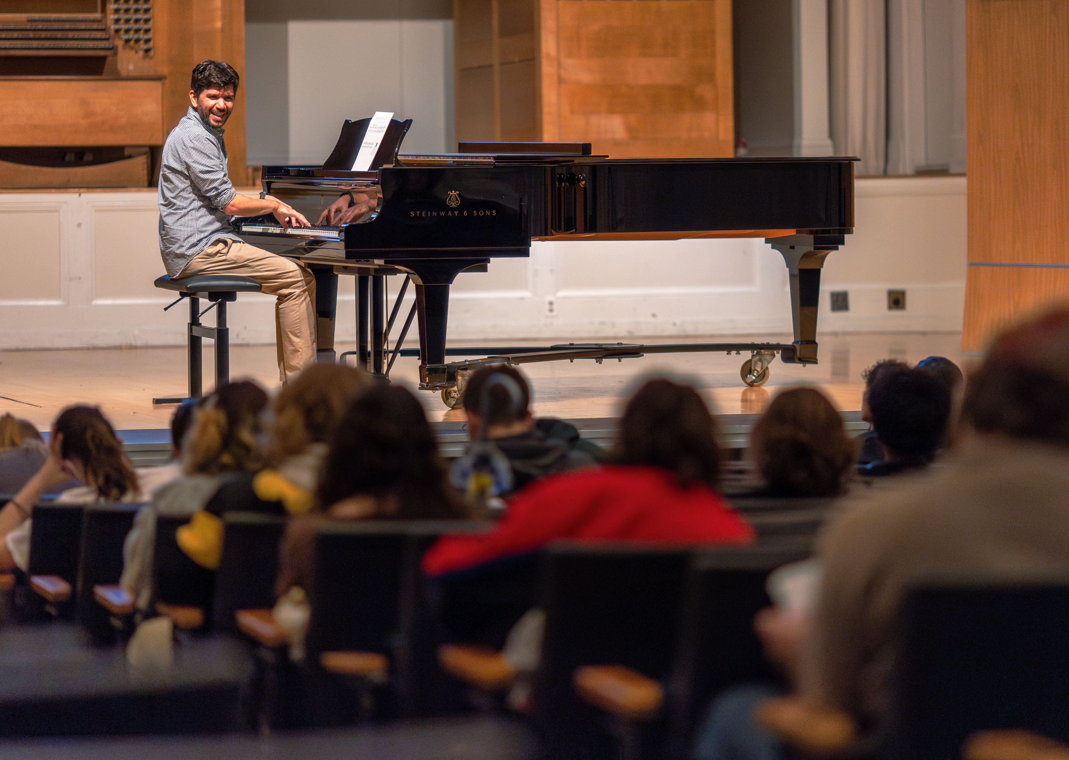 An man sits at a piano on a stage addressing an auditorium of students