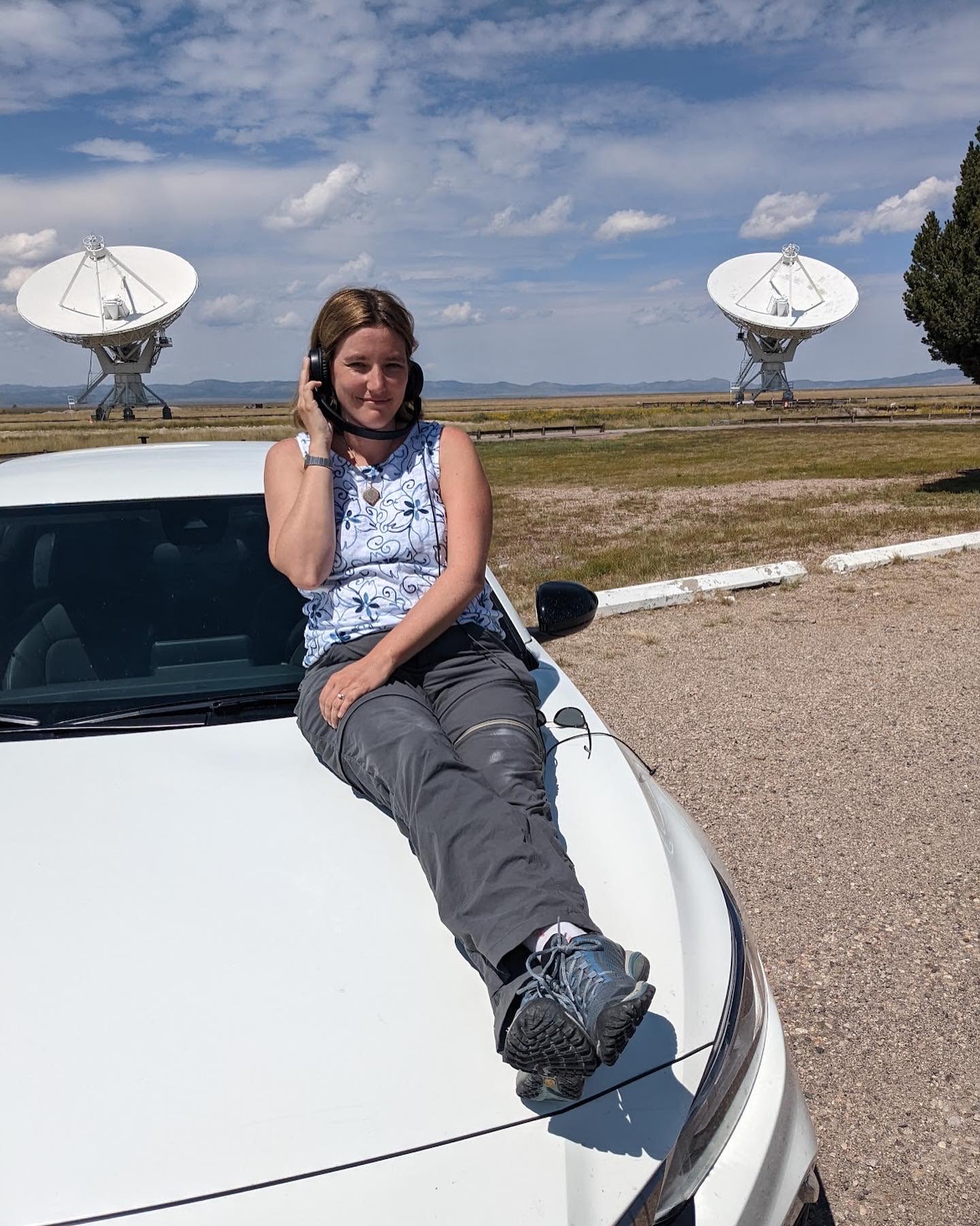Cendes lying on the hood of a car listening to headphones with radio telescope dishes in the background