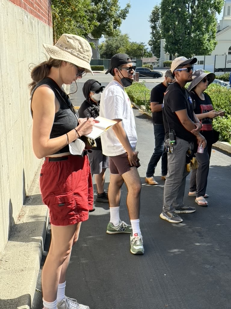 Outside, a person wears a tan bucket hate and red shorts while writing in a notepad