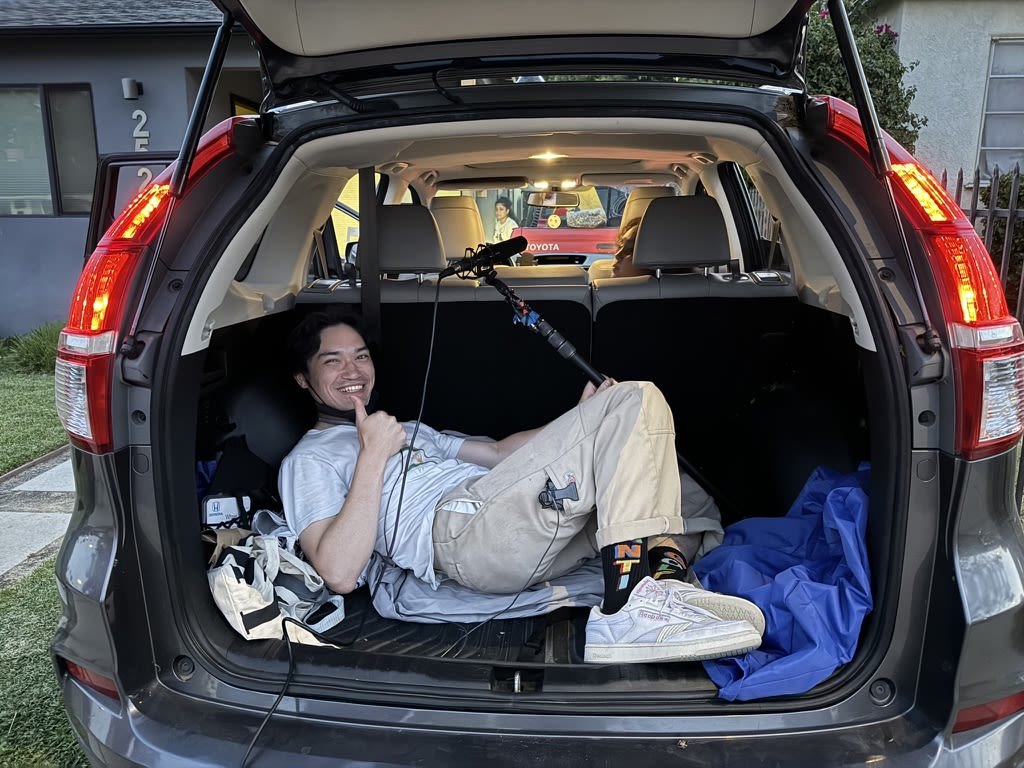 a person lays on his back holding a microphone in his left hand while in the trunk of a car with the hatch open