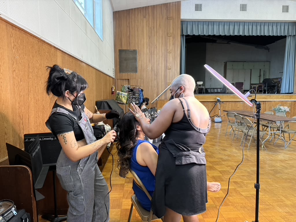 two hair dressers work on an actor in a cafeteria setting